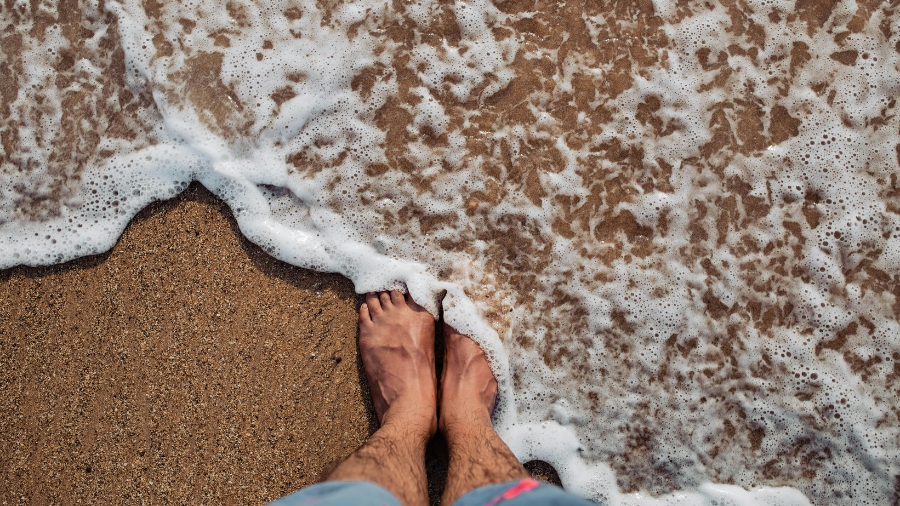 Stopy na plaży otulone falami spienionego morza - zdjęcie poglądowe.